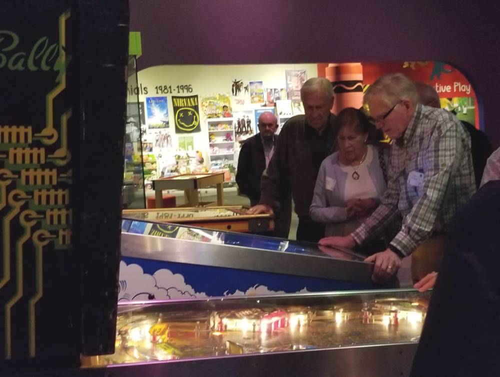 A man stands in front of a pinball machine while another man and woman watch him play